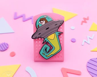 May include: A whimsical enamel pin featuring a brown otter piloting a gray UFO. The pin is set against a pink background with colorful geometric shapes. The pin has a teal, yellow, and green design.