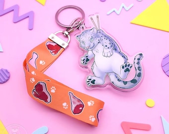 Op de afbeelding: Een heldere acryl sleutelhanger met een cartoon sneeuwluipaard en eetstokjes. Het is bevestigd aan een oranje lanyard bedrukt met vlees en pootafdrukken. De achtergrond is roze met kleurrijke geometrische vormen.