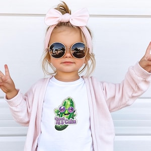 May include: A young child wearing round sunglasses, a pink headband, and a light pink cardigan. The child is wearing a white t-shirt with a cartoon pickle graphic that says "Cool As a Cuke". The child is making a peace sign with both hands.