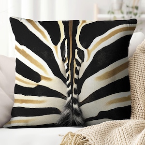 May include: Square throw pillow featuring a close-up of a zebra's rear, showcasing black and white stripes with tan accents. The pillow has a fluffy tail and is placed on a white couch with a beige blanket.