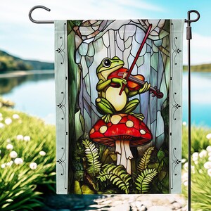 May include: A garden flag featuring a whimsical illustration of a green frog playing a violin while seated on a red mushroom with white spots. The background depicts a stained-glass window effect with a serene lake view.