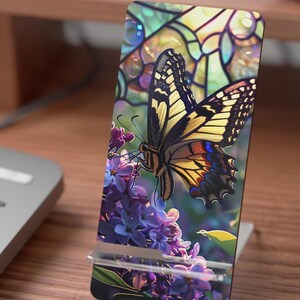 May include: A phone stand featuring a colorful butterfly design with yellow, black, and orange wings, perched on purple flowers. The background has a stained glass effect. The stand is made of clear plastic and sits on a wooden surface.