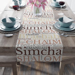 May include: A beige table runner with a repeating pattern of Hebrew words in brown and peach. The words include "Shalom", "Simcha", "L'Chaim", "Brachah", "Tzedakah", "Emanua", "Mitvah", and "Challah".