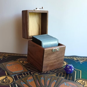 May include: A wooden box with a hinged lid, open to reveal a stack of playing cards. The box is made of dark wood with a light wood interior. The box is sitting on a green and brown patterned surface with a purple die.