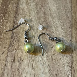 May include: A pair of dangle earrings with a light green pearl-like bead and silver-colored hooks. Two clear earring backs are also present. The earrings are displayed on a wooden surface.
