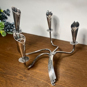 May include: A silver-toned metal candelabra featuring four individual candle holders shaped like calla lily flowers. The holders are connected by curved, slender metal arms that meet at a central point, extending into a decorative base.