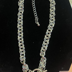 May include: Silver-toned chain necklace with a toggle clasp. The necklace features a series of interlocking oval links, creating a chunky, textured appearance. An adjustable chain extends from the clasp. The necklace is displayed against a black background.