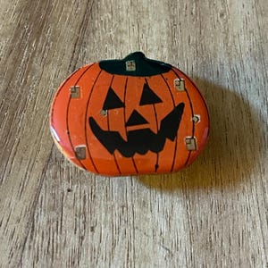 May include: A round, orange Halloween decoration shaped like a jack-o'-lantern. The pumpkin has a black, smiling face and a green stem. Small square accents are placed around the pumpkin's face.