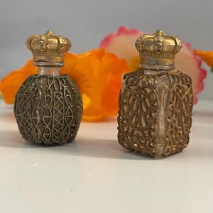 May include: Two antique perfume bottles with ornate metal filigree and crown-shaped stoppers. One bottle is round, the other is square. The bottles are gold-toned and sit on a white surface, with orange and pink flowers in the background.