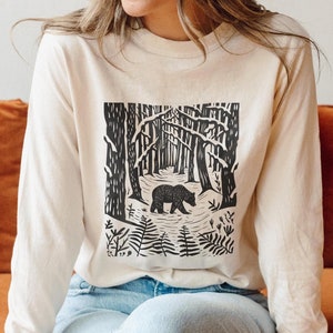 May include: A long-sleeved shirt in ivory with a black and white graphic print of a bear walking through a forest. The graphic features trees, ferns, and a bear.