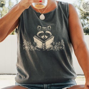 May include: A gray tank top featuring a raccoon reading a book, with the word "PEPPER" at the bottom. The raccoon illustration is in the center, surrounded by leafy sprigs. The tank top is paired with denim shorts.