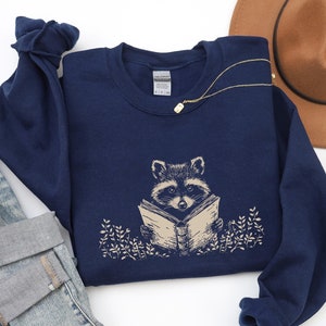 Peut inclure: Sweat-shirt bleu marine avec un dessin beige d'un raton laveur lisant un livre. Le dessin comprend un feuillage beige autour du raton laveur. Le mot "NAVY" est imprimé en bas à droite.