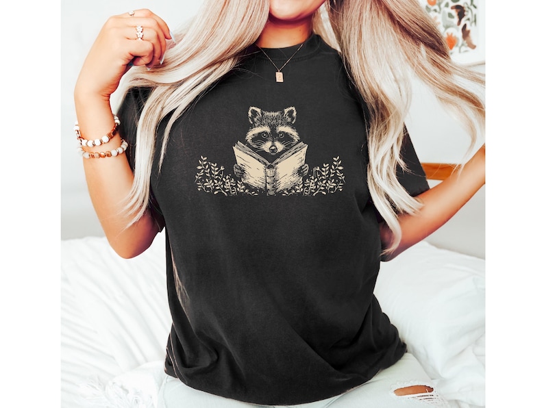 May include: A black t-shirt with a graphic of a raccoon reading a book. The raccoon is sitting in a field of flowers.