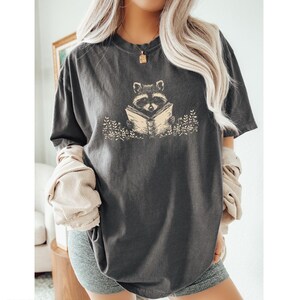 May include: A dark gray t-shirt with a graphic of a raccoon reading a book. The raccoon is surrounded by small flowers.