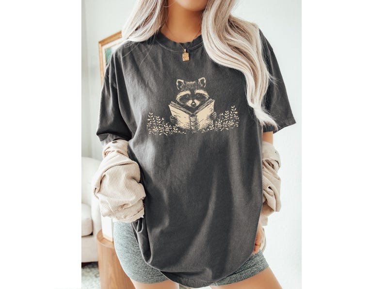 May include: A dark gray t-shirt with a graphic of a raccoon reading a book. The raccoon is surrounded by small flowers.