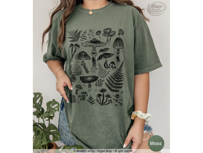 A moss colored Comfort Colors graphic t-shirt featuring a monochromatic array of illustrated botanical mushrooms. It’s a perfect gift for nature lovers, mushroom foragers, granola girls, and forest wanderers.