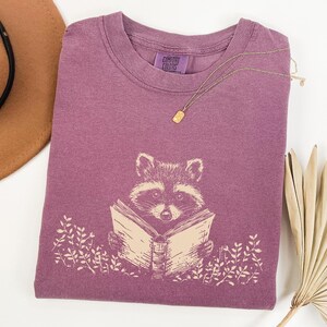 May include: A purple t-shirt with a graphic of a raccoon reading a book. The text "Comfort Colors" is printed on the shirt.