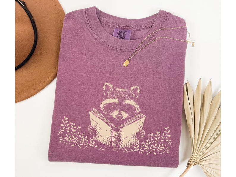 May include: A purple t-shirt with a graphic of a raccoon reading a book. The text "Comfort Colors" is printed on the shirt.