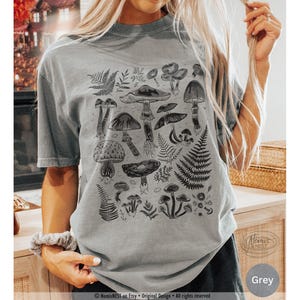 A grey colored Comfort Colors graphic t-shirt featuring a monochromatic array of illustrated botanical mushrooms. It’s a perfect gift for nature lovers, mushroom foragers, granola girls, and forest wanderers.