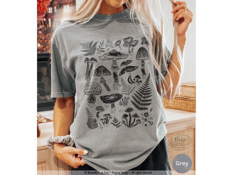 A grey colored Comfort Colors graphic t-shirt featuring a monochromatic array of illustrated botanical mushrooms. It’s a perfect gift for nature lovers, mushroom foragers, granola girls, and forest wanderers.