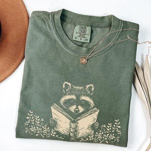 May include: A green t-shirt with a graphic of a raccoon reading a book. The shirt has the text "Comfort Colors" and "S - P - CH" printed on the tag.
