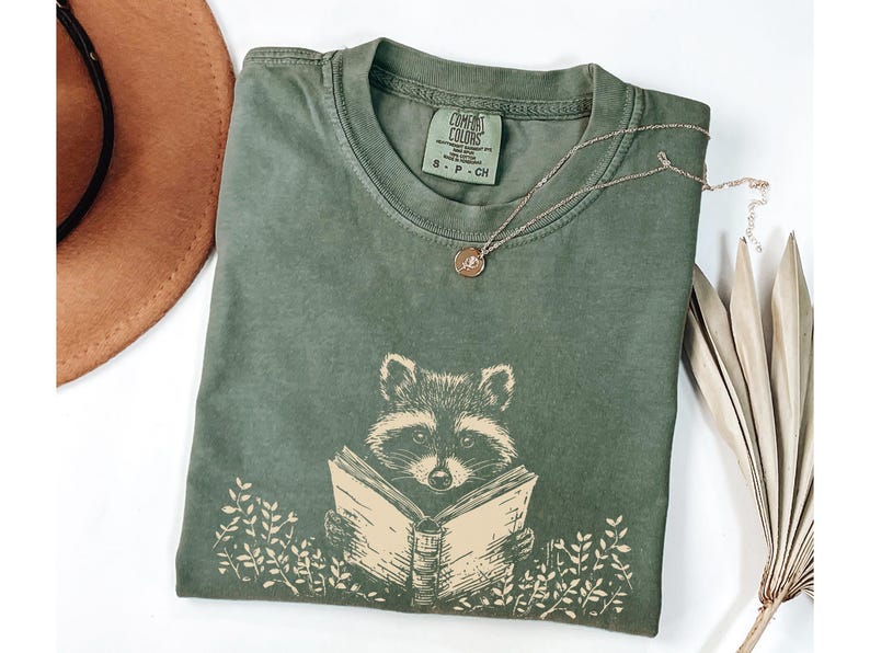 May include: A green t-shirt with a graphic of a raccoon reading a book. The shirt has the text "Comfort Colors" and "S - P - CH" printed on the tag.