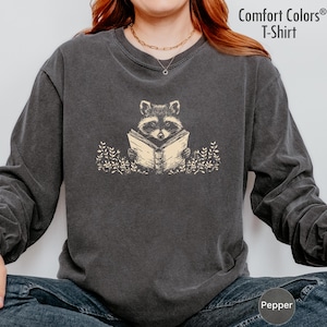 May include: Dark gray Comfort Colors T-shirt with a raccoon reading a book. The raccoon is tan and black, and the book is tan. The shirt has long sleeves. The text "Comfort Colors T-Shirt" is in the upper right corner.
