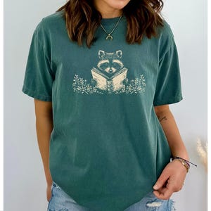 May include: A teal blue t-shirt with a graphic of a raccoon reading a book. The raccoon is surrounded by leaves and flowers.