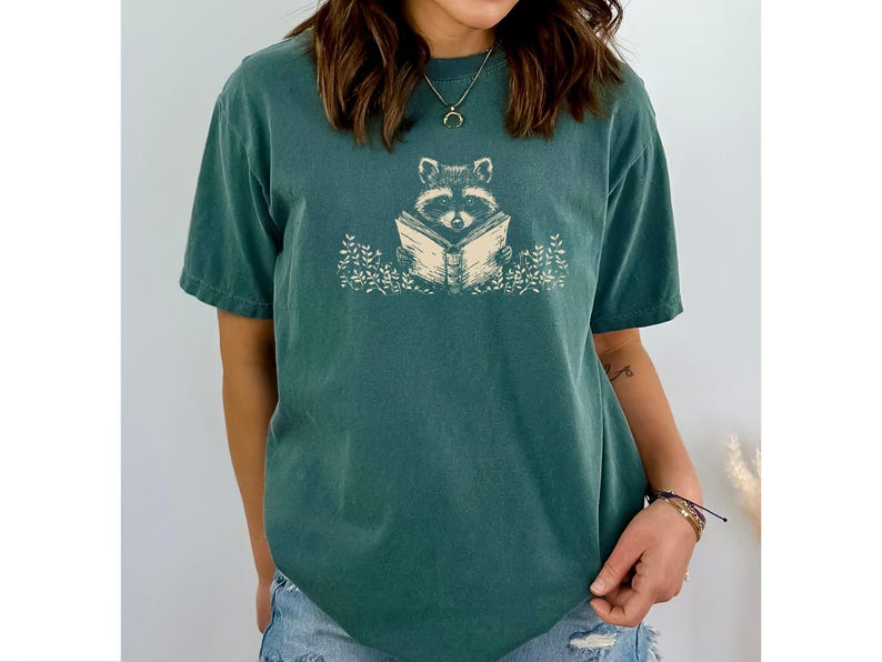 May include: A teal blue t-shirt with a graphic of a raccoon reading a book. The raccoon is surrounded by leaves and flowers.