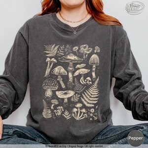 A pepper colored Comfort Colors long sleeve mushroom graphic t-shirt featuring illustrated botanical mushrooms which is perfect for granola girls, mushroom fans, those seeking a forestcore aesthetic, or nature lovers.