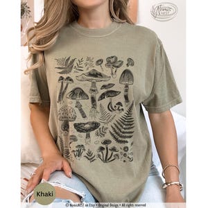 A khaki colored Comfort Colors graphic t-shirt featuring a monochromatic array of illustrated botanical mushrooms. It’s a perfect gift for nature lovers, mushroom foragers, granola girls, and forest wanderers.