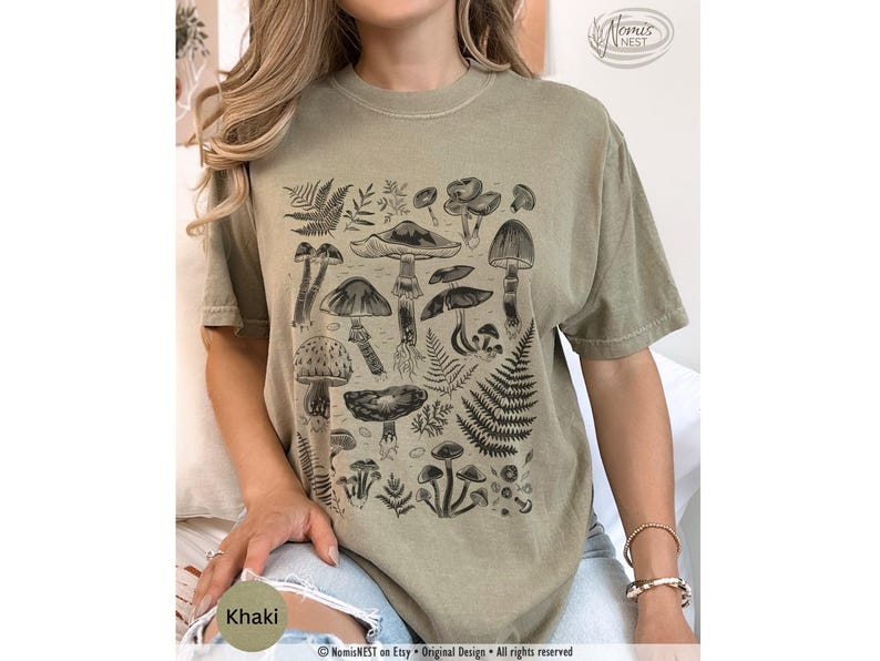 A khaki colored Comfort Colors graphic t-shirt featuring a monochromatic array of illustrated botanical mushrooms. It’s a perfect gift for nature lovers, mushroom foragers, granola girls, and forest wanderers.