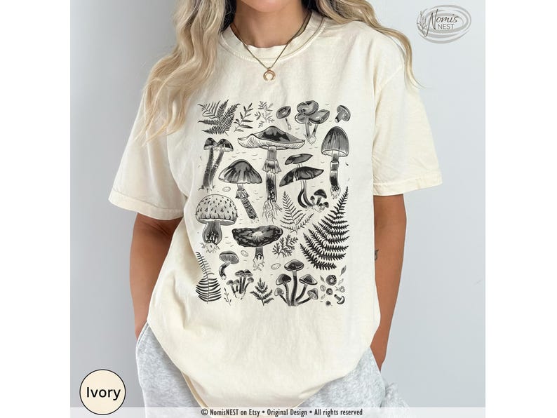 An ivory colored Comfort Colors graphic t-shirt featuring a monochromatic array of illustrated botanical mushrooms. It’s a perfect gift for nature lovers, mushroom foragers, granola girls, and forest wanderers.
