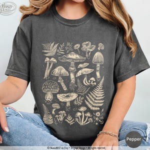 A pepper colored Comfort Colors graphic t-shirt featuring a monochromatic array of illustrated botanical mushrooms. It’s a perfect gift for nature lovers, mushroom foragers, granola girls, and forest wanderers.