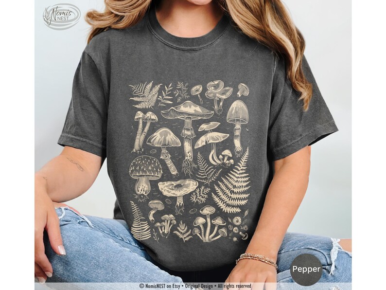A pepper colored Comfort Colors graphic t-shirt featuring a monochromatic array of illustrated botanical mushrooms. It’s a perfect gift for nature lovers, mushroom foragers, granola girls, and forest wanderers.