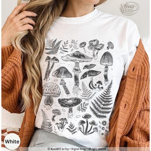 A white colored Comfort Colors graphic t-shirt featuring a monochromatic array of illustrated botanical mushrooms. It’s a perfect gift for nature lovers, mushroom foragers, granola girls, and forest wanderers.