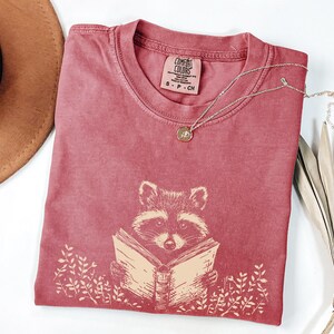 May include: A red t-shirt with a graphic of a raccoon reading a book. The shirt has the text "COMFORT COLORS" and "S - P - CH" printed on the tag.
