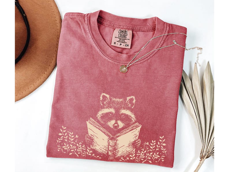 May include: A red t-shirt with a graphic of a raccoon reading a book. The shirt has the text "COMFORT COLORS" and "S - P - CH" printed on the tag.