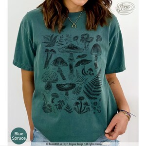 A blue spruce colored Comfort Colors graphic t-shirt featuring a monochromatic array of illustrated botanical mushrooms. It’s a perfect gift for nature lovers, mushroom foragers, granola girls, and forest wanderers.