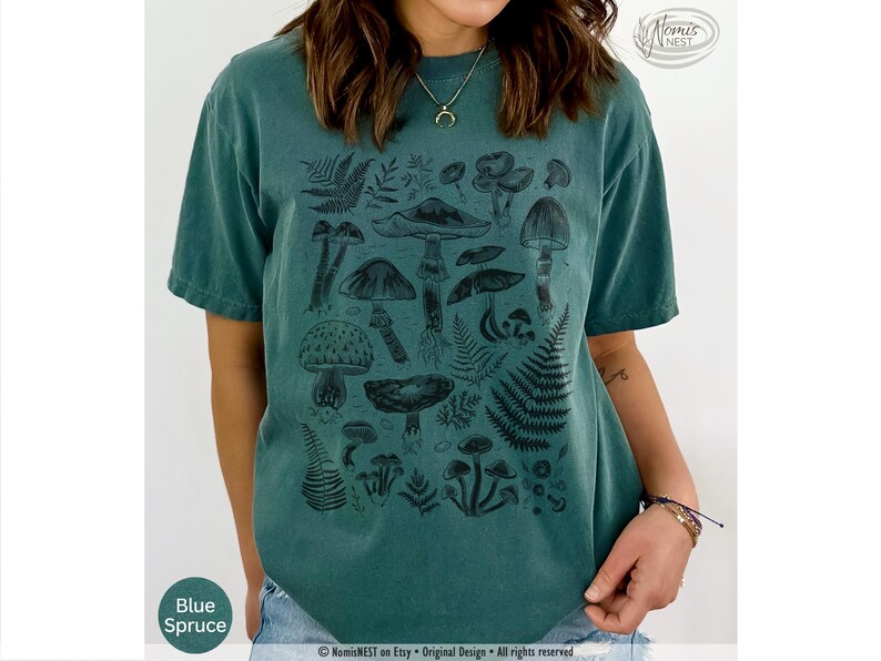 A blue spruce colored Comfort Colors graphic t-shirt featuring a monochromatic array of illustrated botanical mushrooms. It’s a perfect gift for nature lovers, mushroom foragers, granola girls, and forest wanderers.