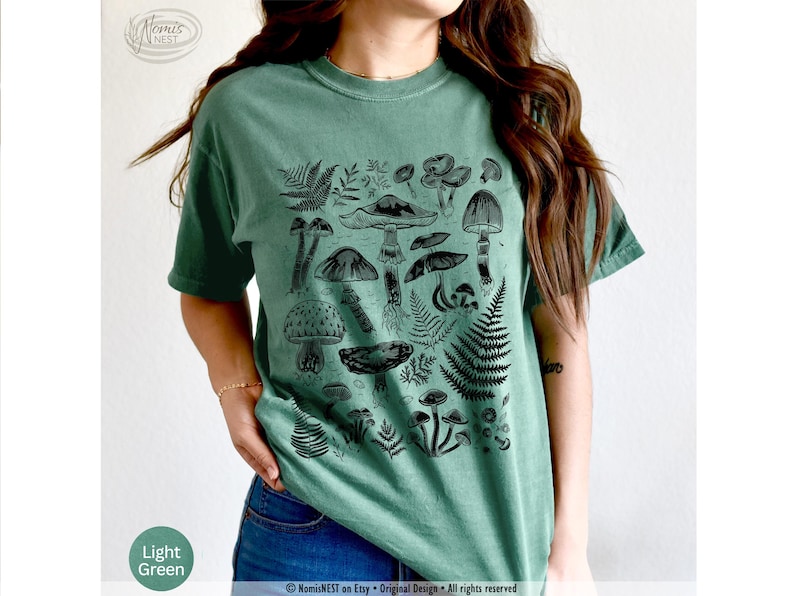 A light green colored Comfort Colors graphic t-shirt featuring a monochromatic array of illustrated botanical mushrooms. It’s a perfect gift for nature lovers, mushroom foragers, granola girls, and forest wanderers.