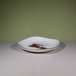 May include: A white ceramic dish with a gold rim and a black and red illustration of a vintage car. The car is labelled "France".