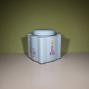 May include: A light blue tin box with a white lid and a colourful illustration of a woman wearing a hat and a dress. The tin box is square shaped with rounded corners.
