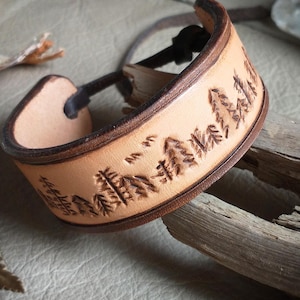 May include: A light brown leather cuff bracelet with a forest scene carved into the surface. The bracelet has a dark brown border and a black cord closure. The design includes trees and birds.
