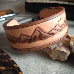 May include: A light brown leather cuff bracelet with a mountain range design. The mountains are etched in a darker brown, creating a contrast against the lighter leather. The bracelet is approximately 1.5 inches wide.