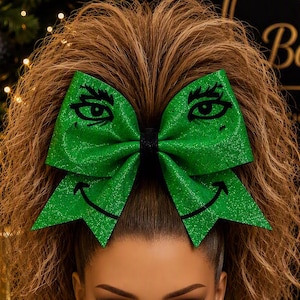 Peut inclure: Un grand nœud pour cheveux vert vif à paillettes avec une impression graphique noire représentant des yeux et une bouche souriante. Le nœud est fixé sur une queue-de-cheval haute de cheveux bruns ondulés. Le tissu a un fini texturé et scintillant.