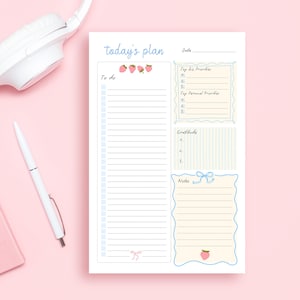 May include: A white notepad with the text "today's plan" in light blue script. The notepad features sections for "To do," "Top Biz Priorities," "Top Personal Priorities," "Gratitude," and "Notes," with strawberry and bow illustrations. A white pen and headphones are in the background.