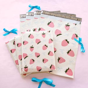 May include: White mailing envelopes and packages with a pink strawberry pattern and light blue bow accents. The strawberries have green stems and are scattered across the surface. The envelopes have a silver closure.