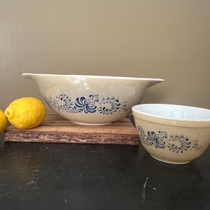May include: Two beige mixing bowls with blue floral designs. One bowl is larger with a spout, and the other is smaller. Two yellow lemons sit on a wooden board next to the bowls.