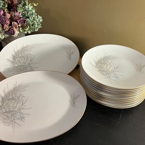 May include: A set of white oval and round ceramic plates with a gray leaf design and gold trim. The plates are stacked on a black surface.